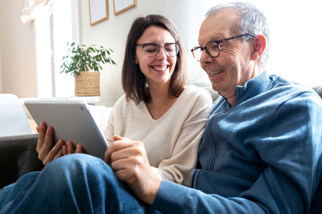 Happy senior man and his young daughter laughing together using digital tablet sitting on sofa at home.